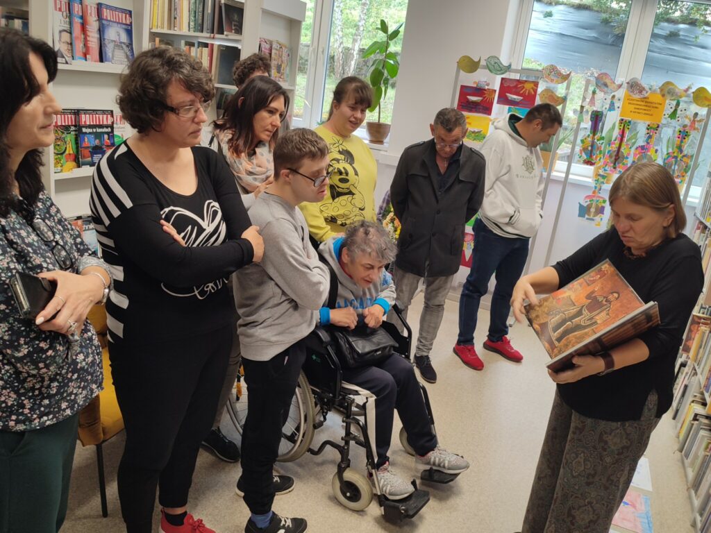 W pomieszczeniu czytelni stoi drupa osób. Jedna z pań prezentuje książkę. Reszta przygląda się.