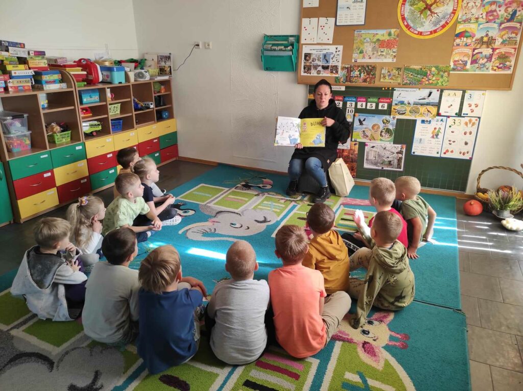 na zdjęciu widać salę przedszkolną, dzieci siedzą w kole na dywanie, bibliotekarz czyta książkę