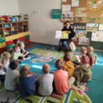 na zdjęciu widać salę przedszkolną, dzieci siedzą w kole na dywanie, bibliotekarz czyta książkę
