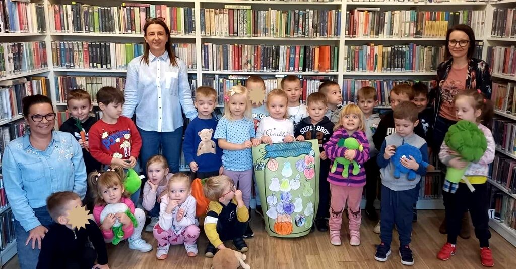 na zdjęciu widać dzieci z trzema dorodłymi kobietami stojące na tle regału z książkami.