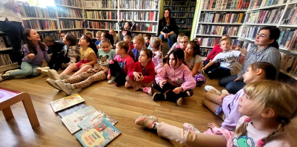 na zdjęciu widać dzieci w sali bibliotecznej, siedzące na podłodze a przed nimi książki