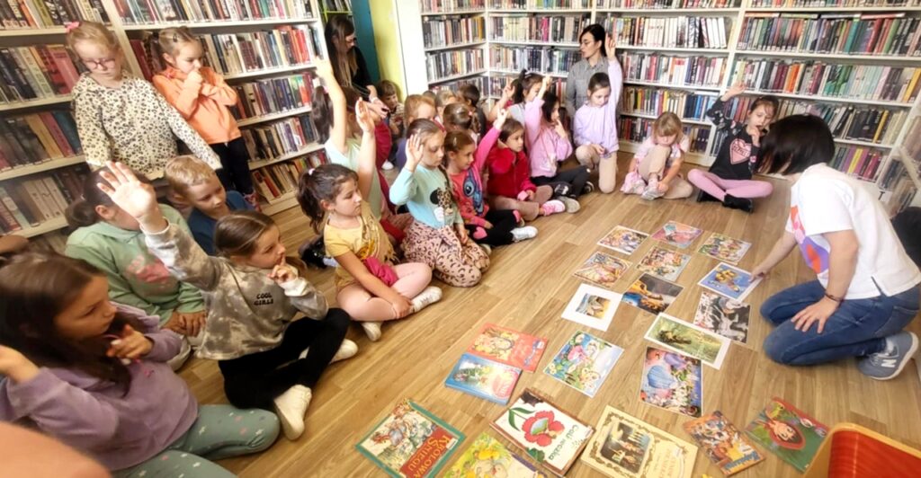 na zdjęciu widać dzieci w sali bibliotecznej, siedzące na podłodze a przed nimi kolorowe ilustracje