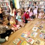 na zdjęciu widać dzieci w sali bibliotecznej, siedzące na podłodze a przed nimi kolorowe ilustracje