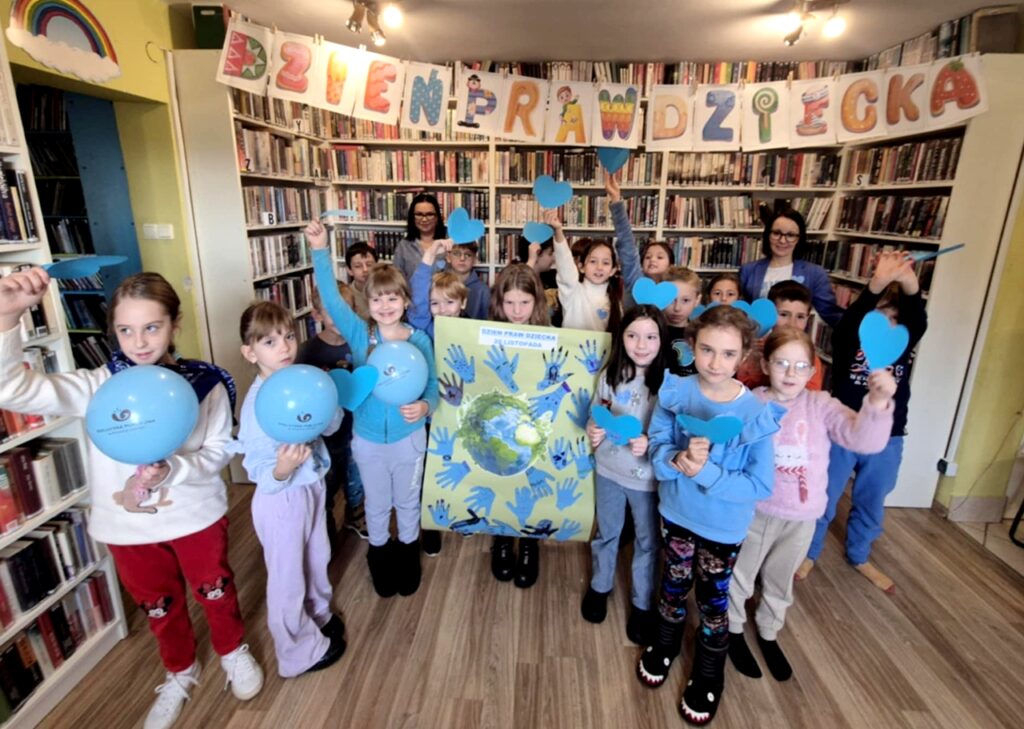 na zdjęciu widać dzieci w sali bibliotecznej trzymające rysunki