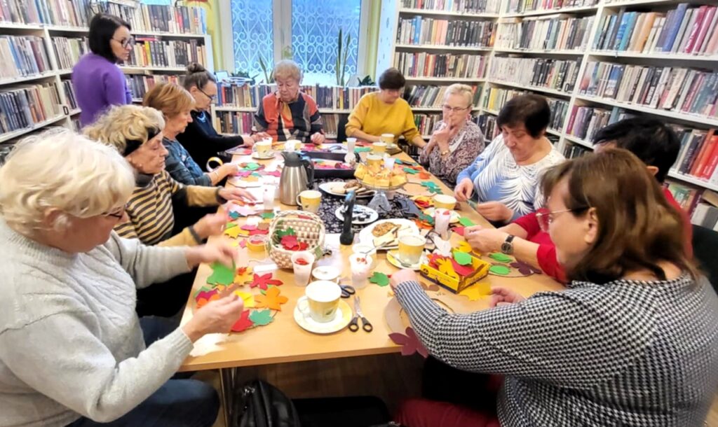 na zdjęciu widać panie siedzące przy stole w sali bibliotecznej i wykonujące prace plastyczne