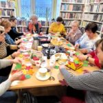 na zdjęciu widać panie siedzące przy stole w sali bibliotecznej i wykonujące prace plastyczne