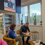 Przy stoliku siedi Jędrzej Pasierski i podpisuję swoją książkę czytelniczce, ktora siedzi obok w fotelu