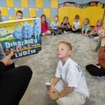 na zdjęciu widać salę przedszkolną, na dywanie siedzą dzieci, bibliotekarz czyta książkę