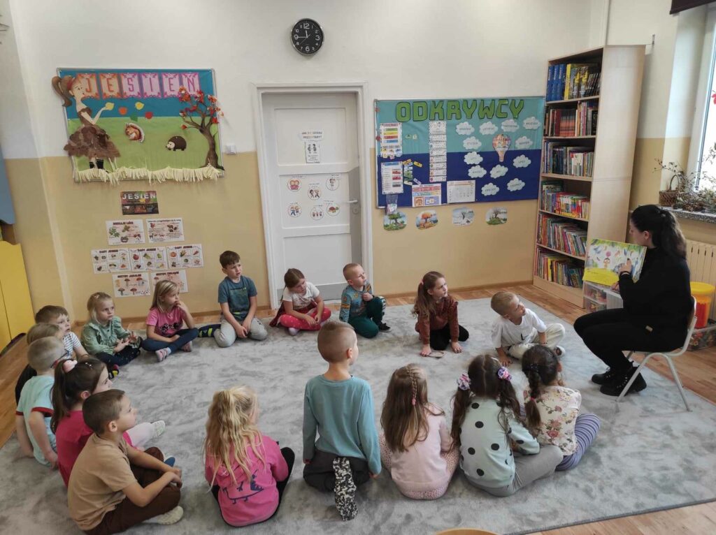 na zdjęciu widać salę przedszkolną, na dywanie siedzą dzieci, bibliotekarz czyta książkę