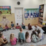 na zdjęciu widać salę przedszkolną, na dywanie siedzą dzieci, bibliotekarz czyta książkę