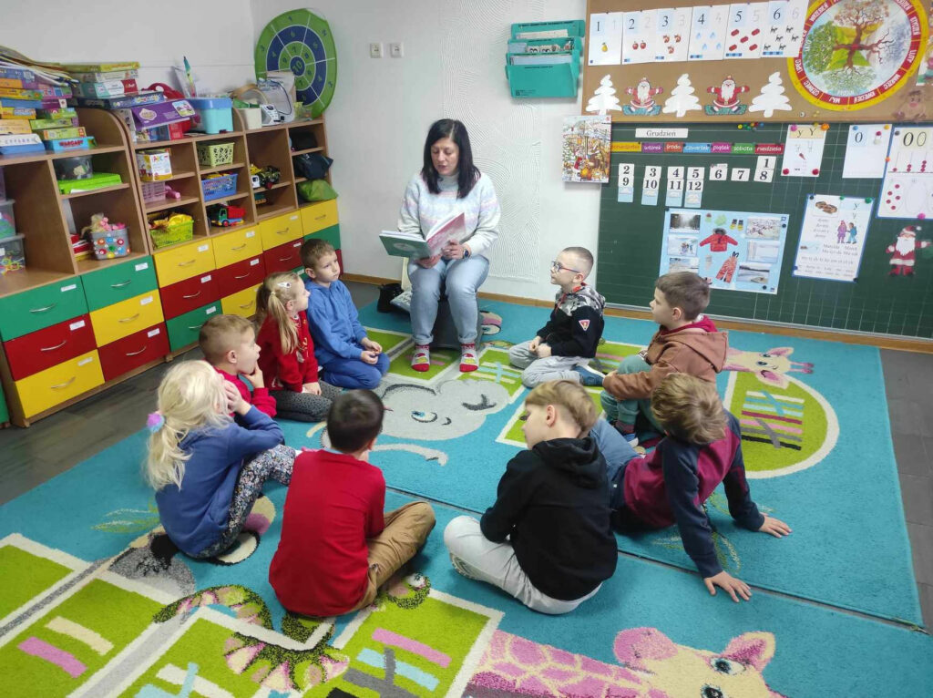 na zdjęciu widać salę przedszkolną, w kole na dywanie siedzą dzieci, bibliotekarz czyta książkę