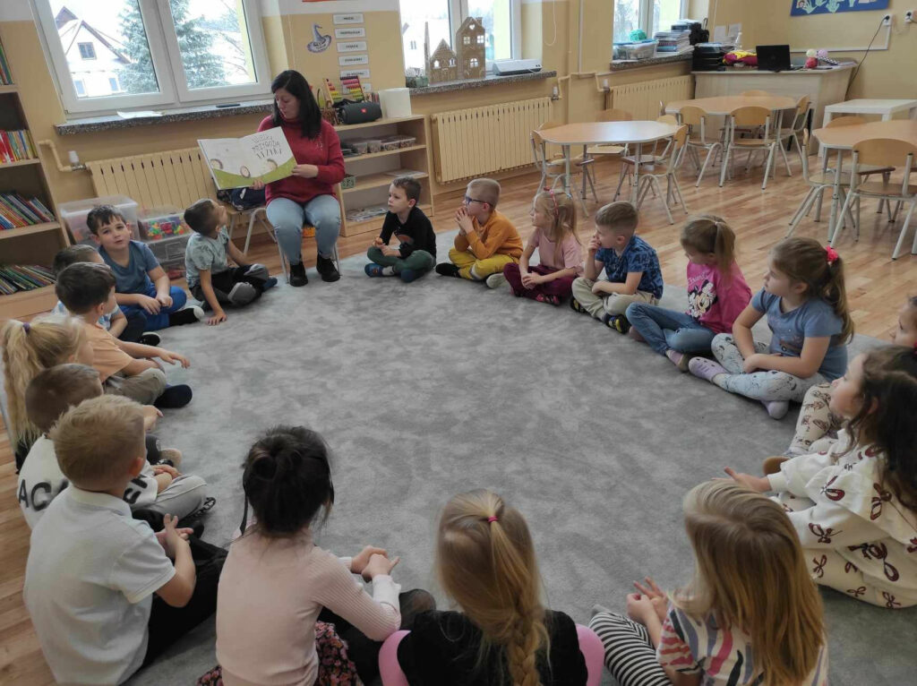 na zdjęciu widać salę przedszkolną, dzieci siedzą w kole na dywanie, bibliotekarz czyta książkę