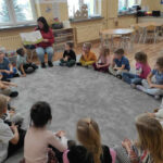 na zdjęciu widać salę przedszkolną, dzieci siedzą w kole na dywanie, bibliotekarz czyta książkę