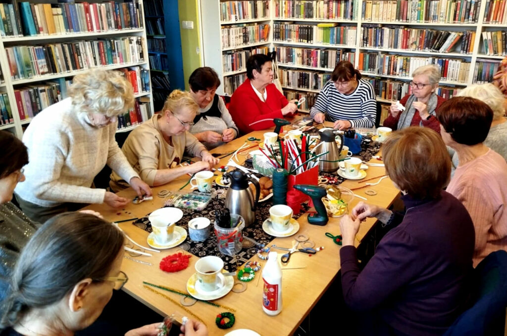 na zdjęciu widać seniorki siedzące wokół stołu w sali bibliotecznej i wykonujące ozdoby świąteczne