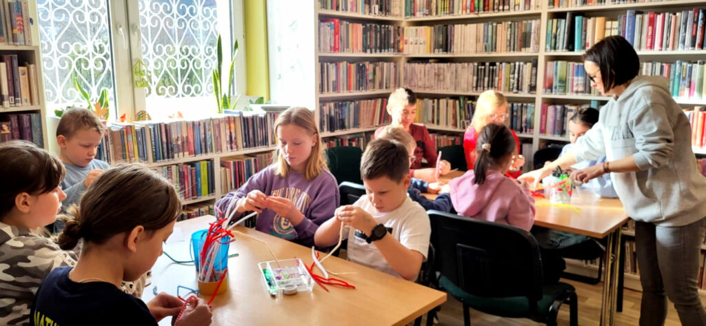 na zdjęciu widać dzieci siedzące przy stołach w sali bibliotecznej i wykonujące prace