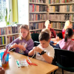 na zdjęciu widać dzieci siedzące przy stołach w sali bibliotecznej i wykonujące prace