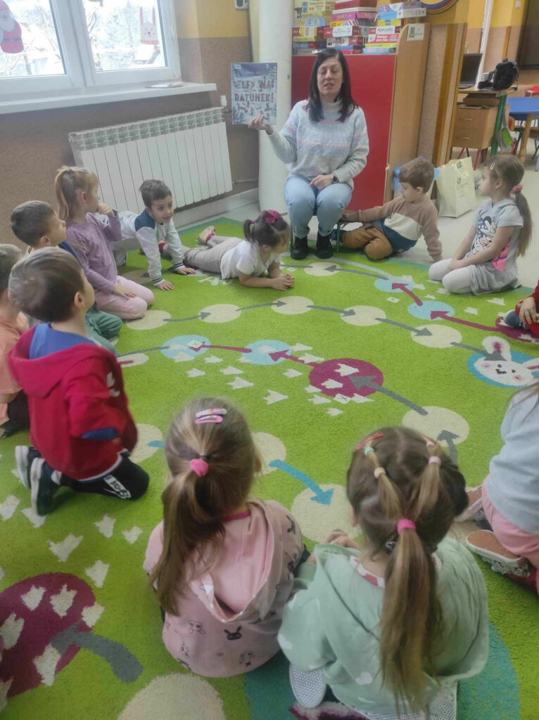 na zdjęciu widać salę przedszkolną, na dywanie w kole siedzą dzieci, bibliotekarz czyta książkę