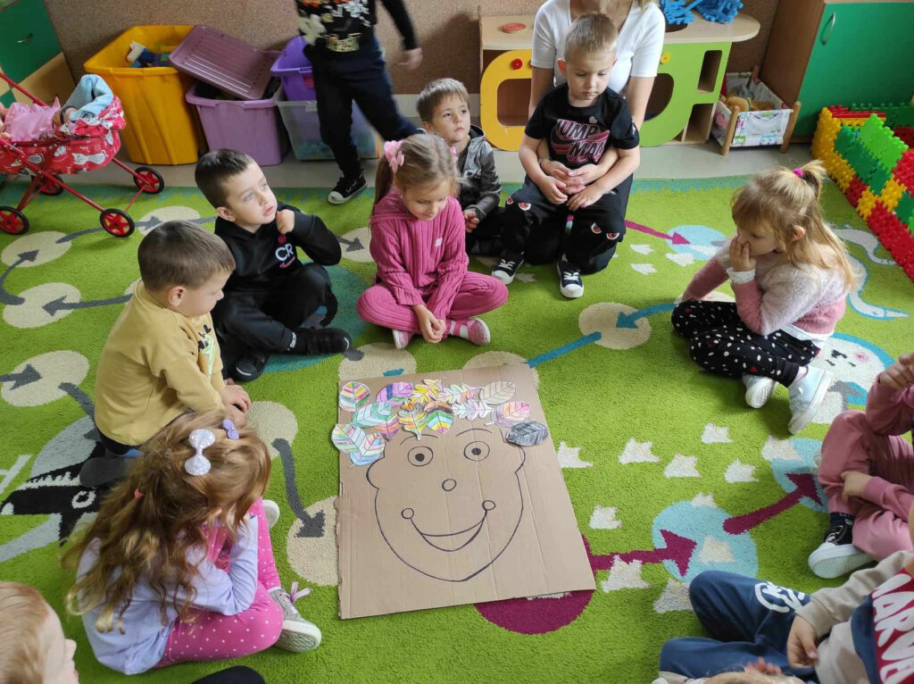 na zdjęciu widać salę przedszkolną, dzieci siedzą na dywanie, biorą udział w zabawie