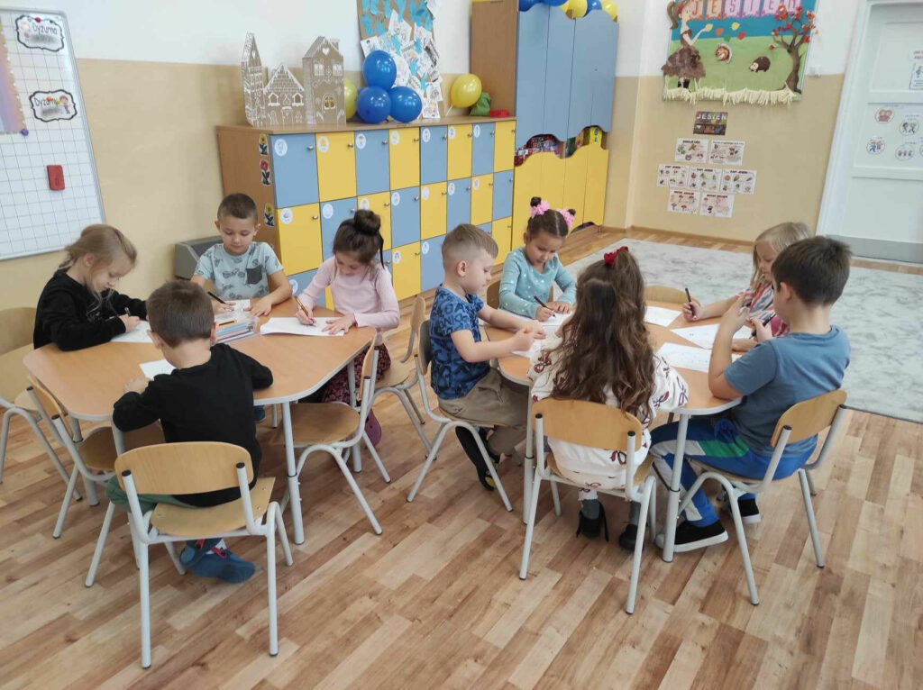 na zdjęciu widać salę przedszkolną, dzieci siedzą przy stolikach, wykonują pracę plastyczną