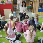 a zdjęciu widać salę przedszkolną, dzieci siedzą na dywanie, bibliotekarz czyta książkę