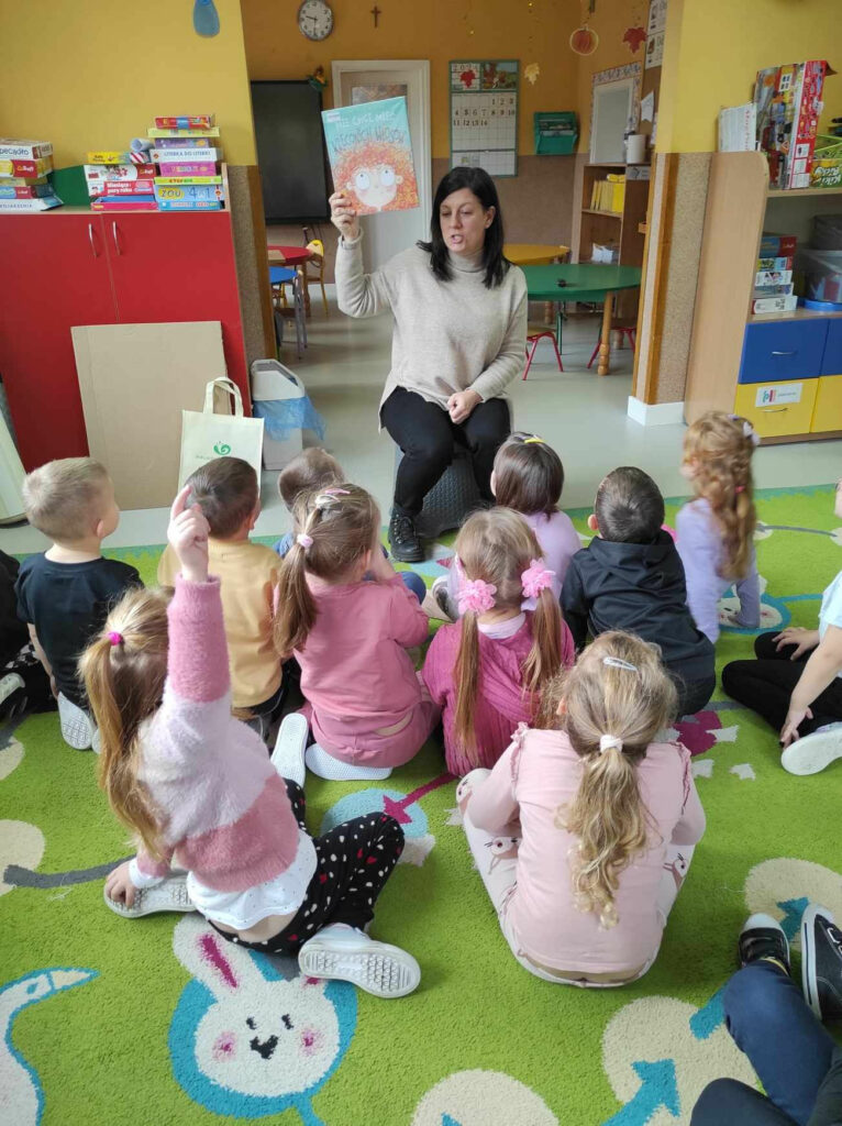 a zdjęciu widać salę przedszkolną, dzieci siedzą na dywanie, bibliotekarz czyta książkę