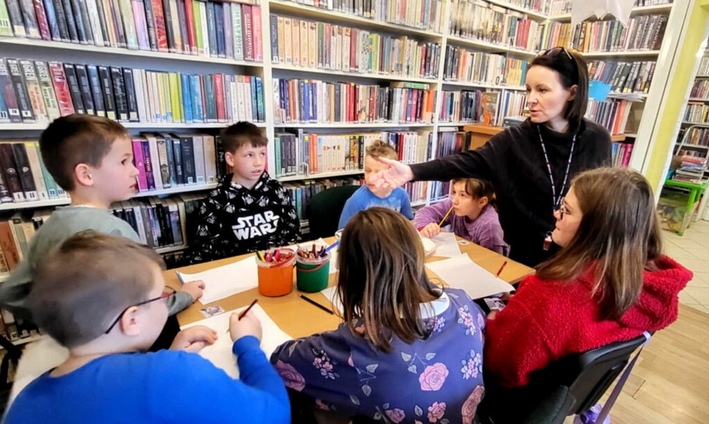 na zdjęciu widać dzieci siedzące wokół stołu w sali bibliotecznej
