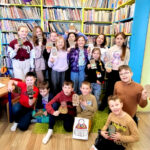 na zdjęciu widać dzieci w sali bibliotecznej, stoją na tle regału. Prezentują swoje prace.