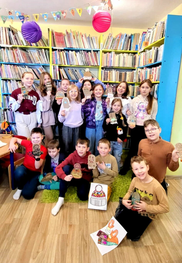 na zdjęciu widać dzieci w sali bibliotecznej, stoją na tle regału. Prezentują swoje prace.
