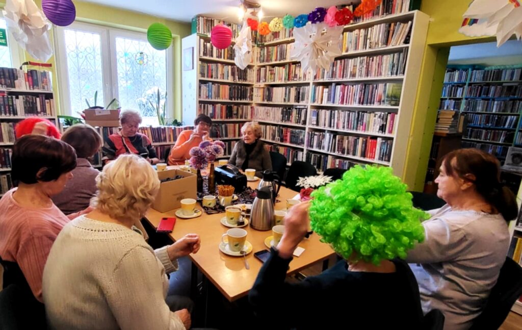 na zdjęciu widać seniorki siedzące wokół stołu w sali bibliotecznej