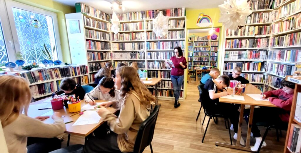 na zdjęciu widać dzieci siedzące przy stołach w sali bibliotecznej i wykonują prace plastyczne.