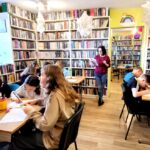 na zdjęciu widać dzieci siedzące przy stołach w sali bibliotecznej i wykonują prace plastyczne.