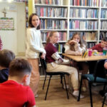 na zdjęciu widać dzieci siedzące przy stołach w sali bibliotecznej