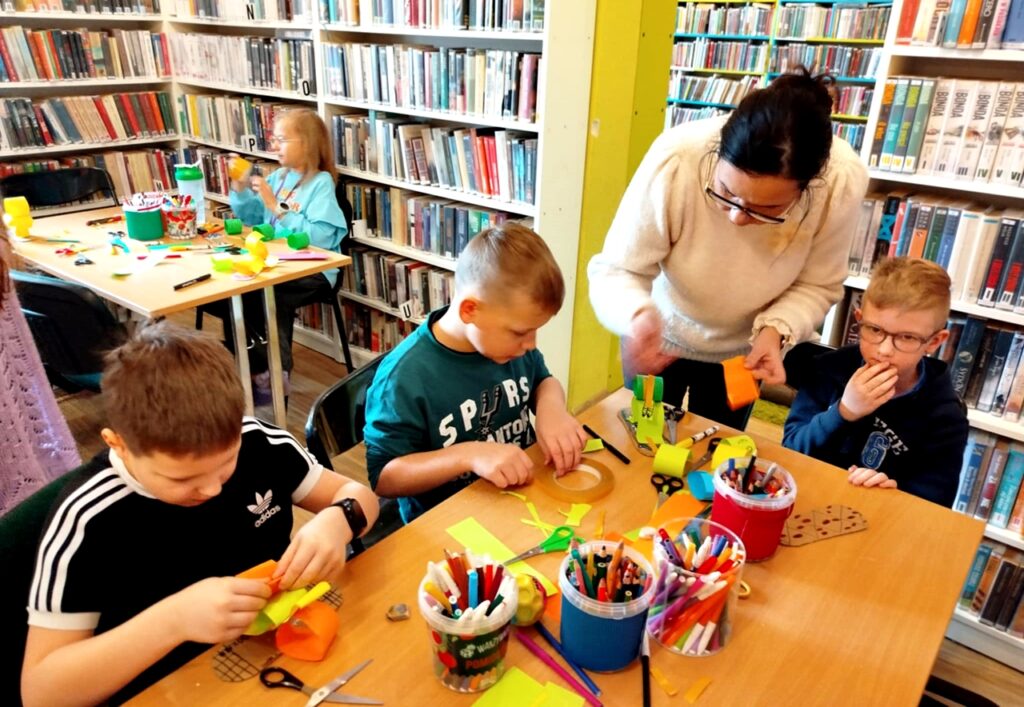 na zdjęciu widać dzieci siedzące przy stołach w sali bibliotecznej, wykonują prace plastyczne.