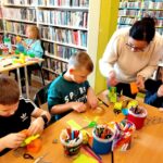 na zdjęciu widać dzieci siedzące przy stołach w sali bibliotecznej, wykonują prace plastyczne.