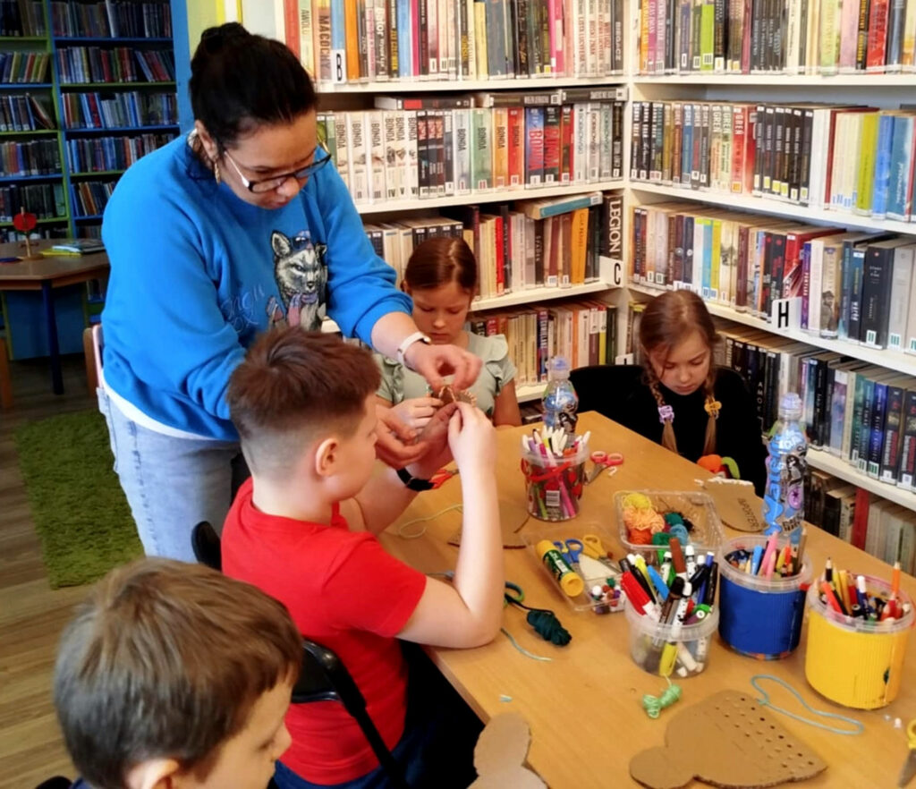 na zdjęciu widać dzieci siedzące przy stołach w sali bibliotecznej