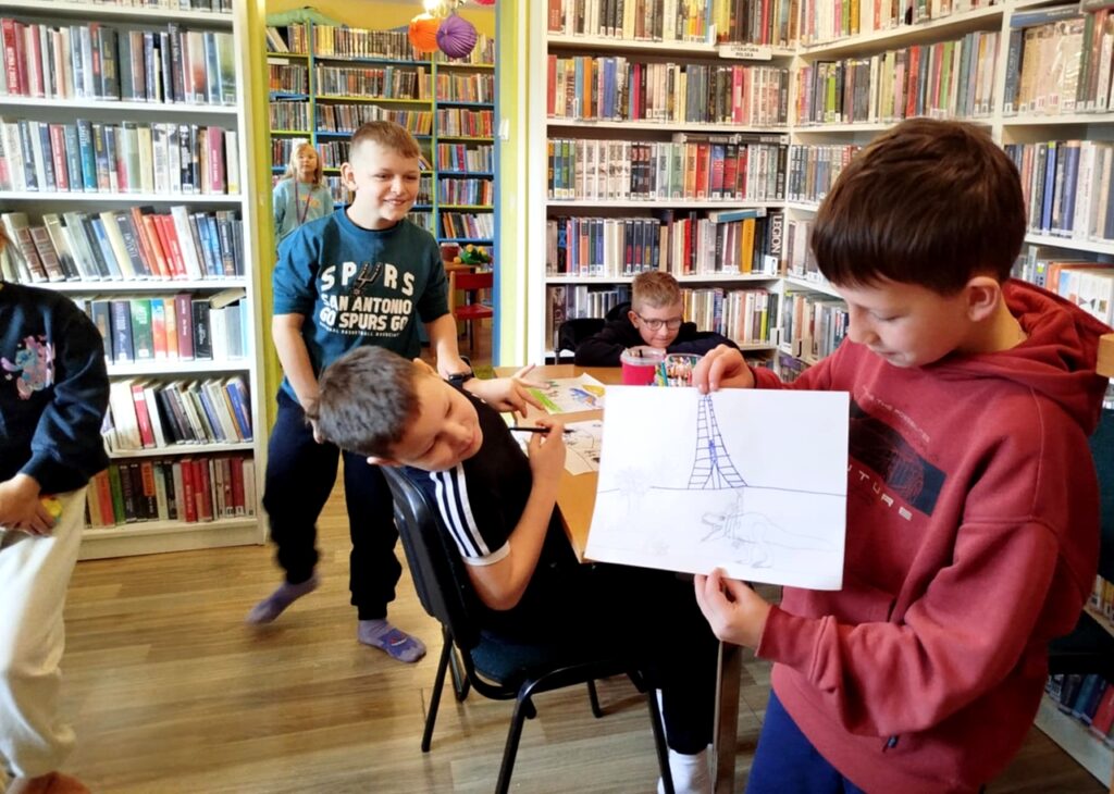 na zdjęciu widać dzieci w bibliotece. Chłopiec prezentuje swoją pracę.