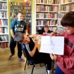 na zdjęciu widać dzieci w bibliotece. Chłopiec prezentuje swoją pracę.