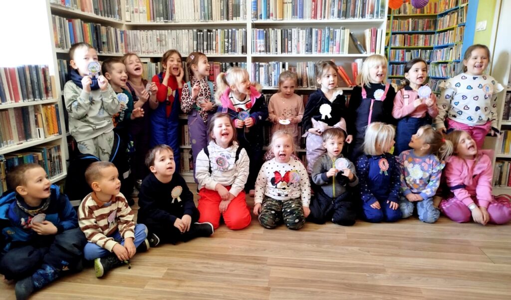 na zdjęciu widać dzieci w sali bibliotecznej stojące na tle regałów z książkami
