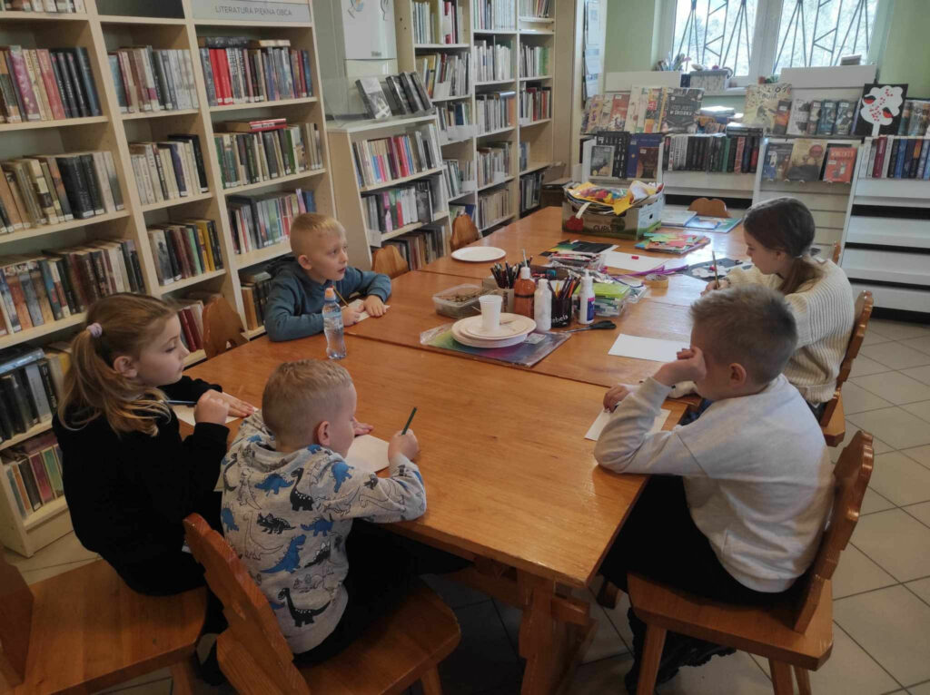 na zdjęciu widać salę biblioteczną, przy stole siedzą dzieci, biorą udział w zabawie