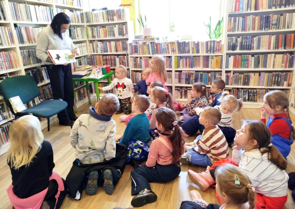 na zdjęciu widać dzieci siedzące na podłodze i bibliotekarkę pokazującą im książkę