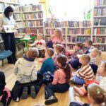 na zdjęciu widać dzieci siedzące na podłodze i bibliotekarkę pokazującą im książkę