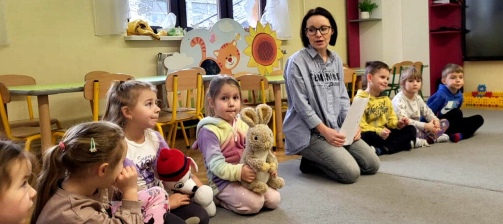na zdjęciu widać dzieci siedzące na podłodze wraz z pana bibliotekarką