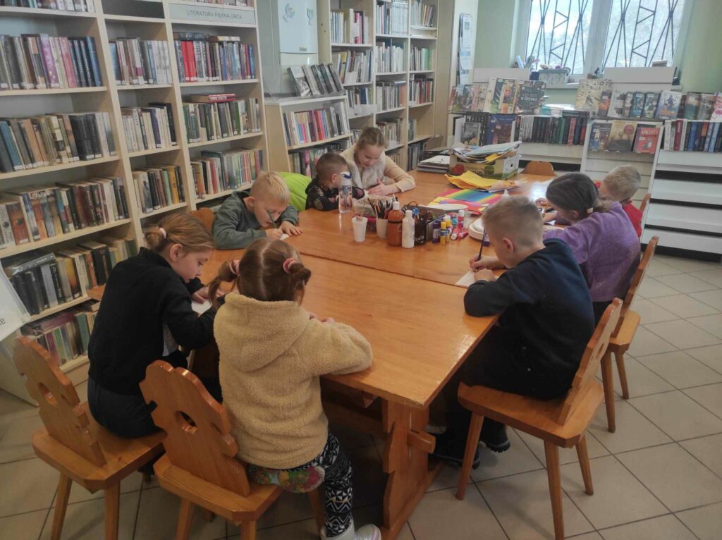 na zdjęciu widać salę biblioteczną, dzieci siedzą przy stole, rozwiązują zadanie