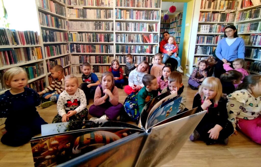 na zdjęciu widać dzieci siedzące na podłodze i bibliotekarkę pokazującą im książkę