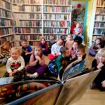 na zdjęciu widać dzieci siedzące na podłodze i bibliotekarkę pokazującą im książkę