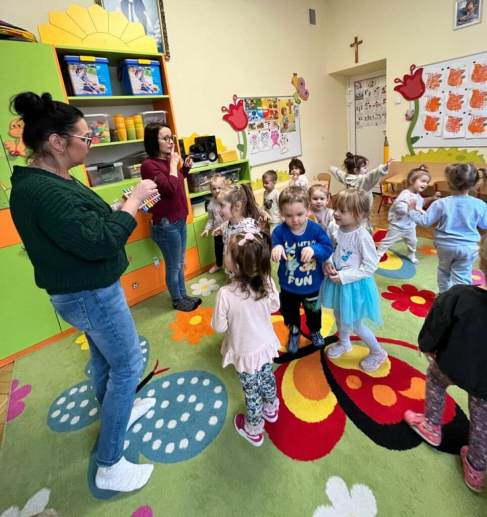 na zdjęciu widać salę przedszkolną, kolorowy dywan oraz dzieci i dwie kobiety wszyscy uczestnicza w zabawie.