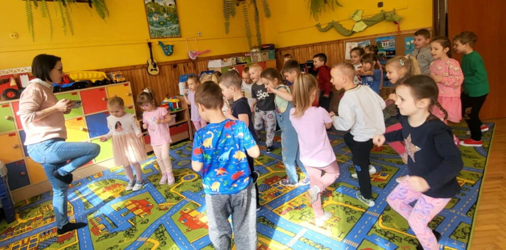 na zdjęciu widać dzieci stojące na jednej nodze w sali przedszkolnej
