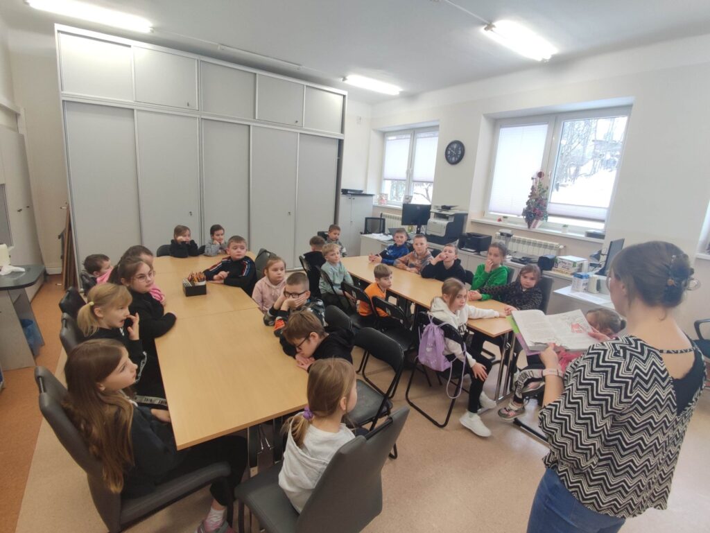 Przy stołach siedzą dwadzieścia pięć dzieci w wieku szkolnym. Pani bibliotekarka czyta im książkę a dzieci słuchają.