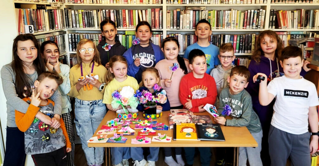 Na zdjęciu stół na którym są prace plastyczne. Za stołem na tle regałów z książkami czternaście dzieci biorących udział w zajęciach bibliotecznych.