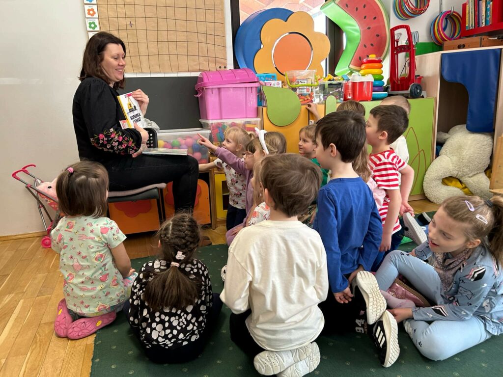 Na krześle w sali przedszkolnej sedzi bibliotekarka, która trzyma w ręku książkę i pokazuje dzieciom. Przedzkolaki w różnokolorowych ubrankach siedzą na dywanie i słuchają pani. W sali widać zabawki różowa skrzynię, klocki, kulki i inne zabawki.